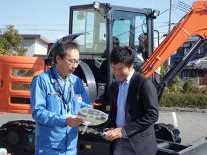 Using the environmental PR uchiwa fan to explain the mini excavator to Mayor Iwanaga (right)