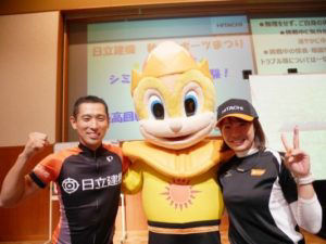 Company employee and para-cyclist Masaki Fujita (left) and trap shooter Yukie Nakayama (right), who both livened up the sports exhibit, strike a pose with Rey-kun (center)